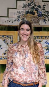 Una mujer de pelo largo y rubio, vestida con una blusa de flores, sonríe delante de una colorida pared de azulejos decorada con motivos florales y geométricos.